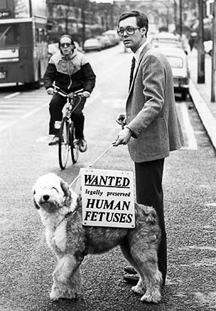 Black and white photo of Daisy the dog carrying the sign on her back and Rick Gibson on a south London street