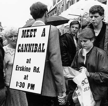 Black and white photo of Gibson entering a crowd of shoppers wearing his sign on a rainy day