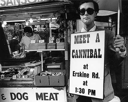 Black and white photo of Gibson posing next to a sign on a market stall which reads DOG MEAT