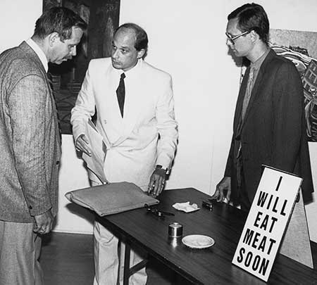 Black and white photo of two police detectives and Gibson standing next to a table with food, cooking utensils and a sign reading "I will eat meat soon"