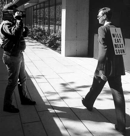 Black and white photo of a police cameraman video recording Gibson's walk