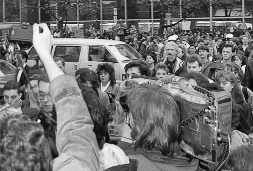 Black and white photo of Gibson surrounded by journalists and a large crowd at the northwest corner of Robson and Burrard