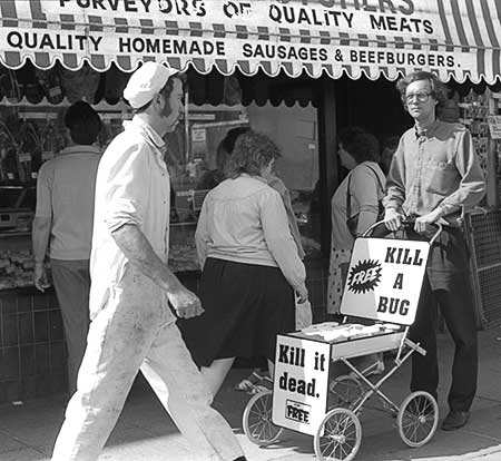 PICTURE: black and white photo of Gibson pushing the pram through central Plymouth.  In this photo Gibson is in front of a butcher shop with a sign reading - Quality homemade sausages and beefburgers