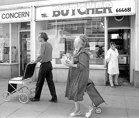 PICTURE: black and white photo of Gibson walking in front another butcher shop.  Store signage reads - Concern...Tel Butcher 664681.  Ms. Piddington is approaching Gibson from behind
