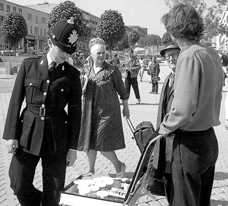 PICTURE: black and white photo of a policeman looking at Gibson's display while Ms. Piddington and a pedestrian watch.